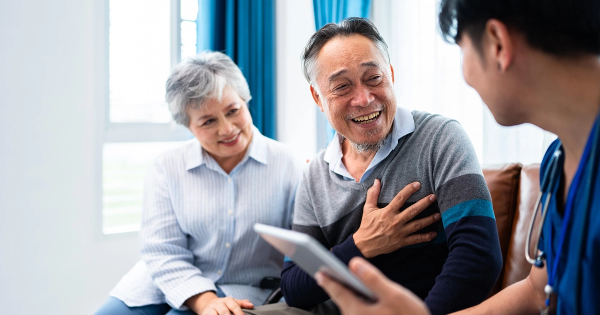 A happy elderly Asian couple smiles at a medical professional holding a tablet during Assisted Living in Charlottesville, VA.
